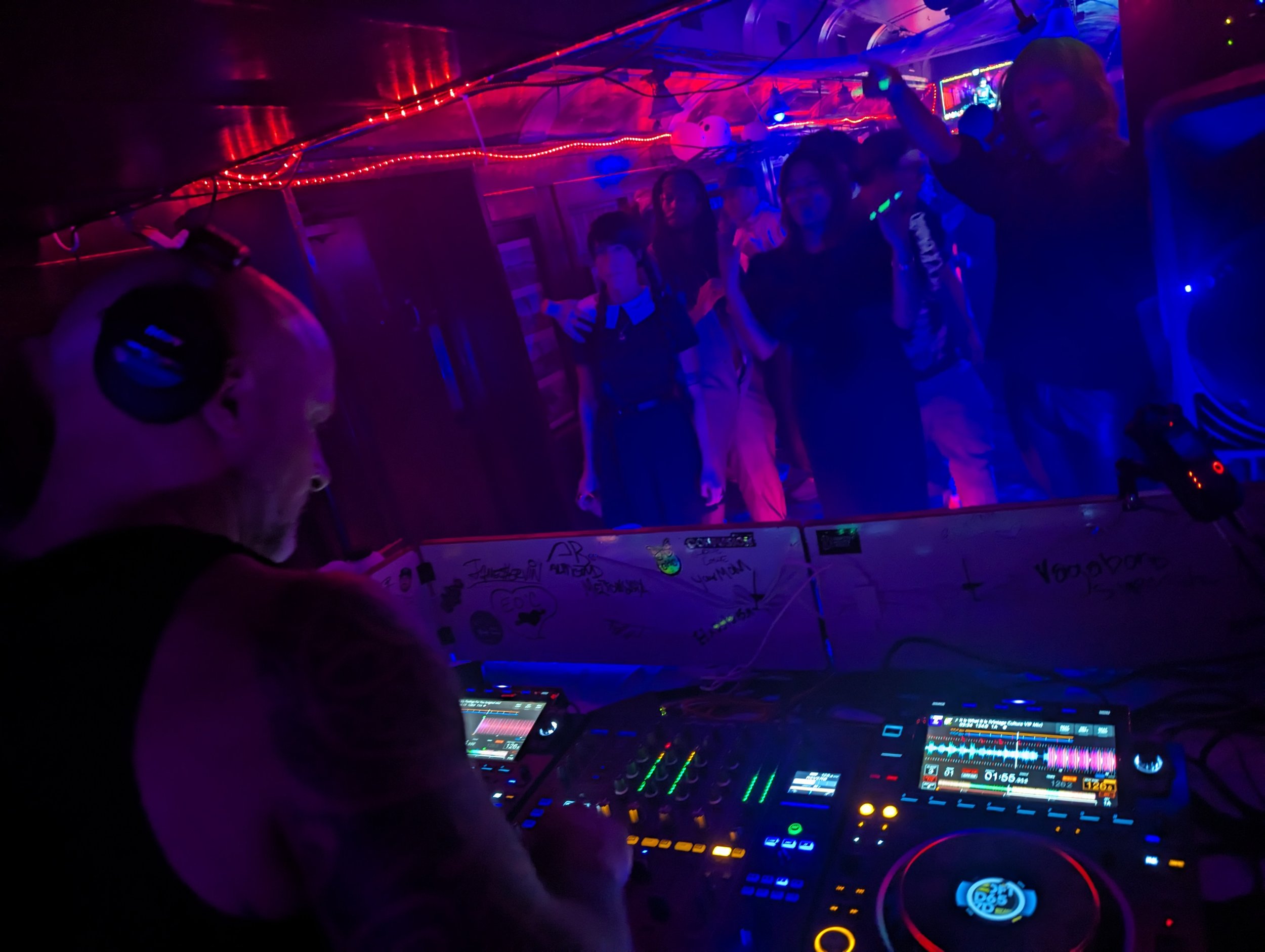 Phil Anthony performing at a nightclub with a crowd visible in the background, illuminated by red and blue lighting effects.
