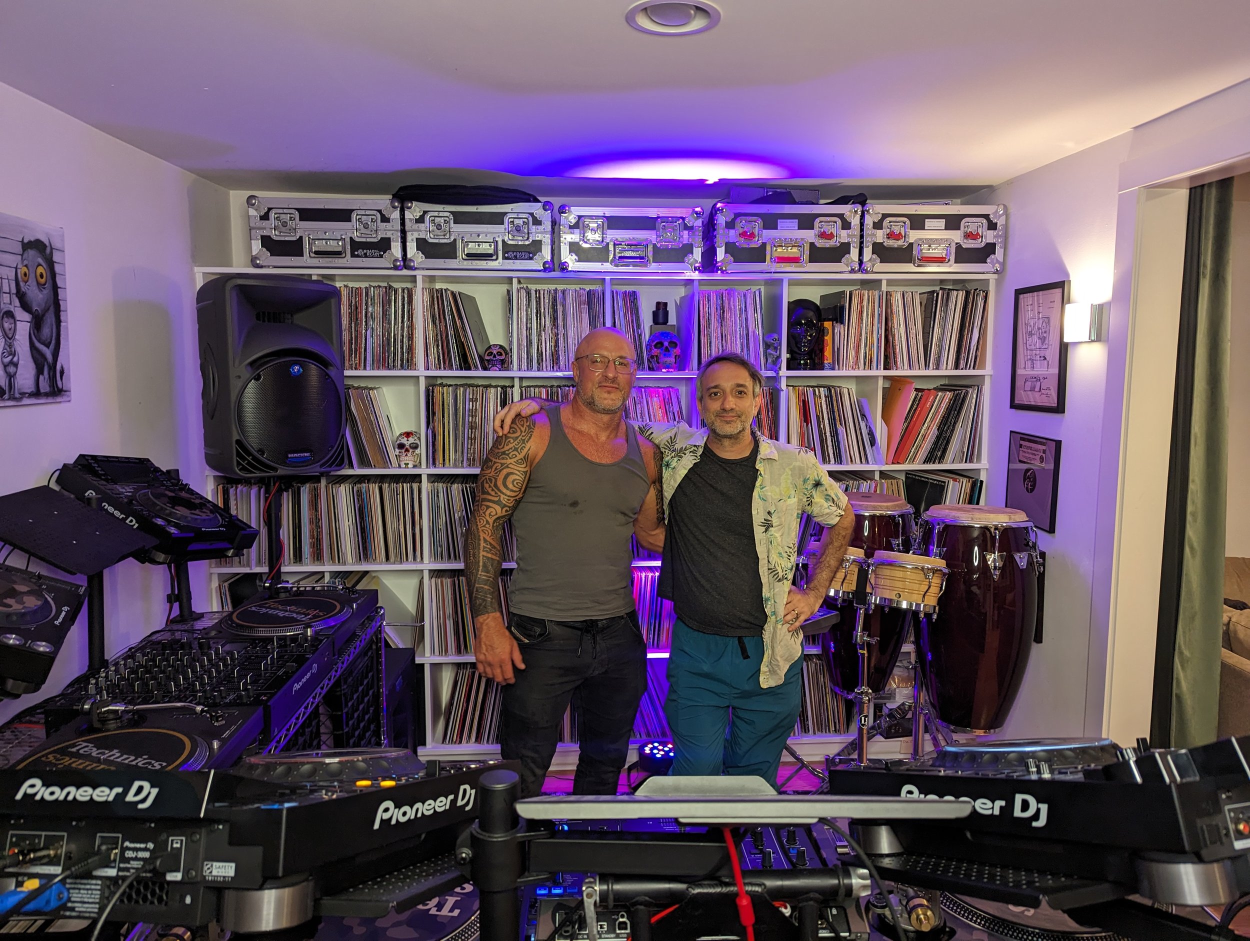 Phil Anthony and Pezzner posing behind Pioneer DJ equipment in a home studio with extensive vinyl record collection and purple ambient lighting.