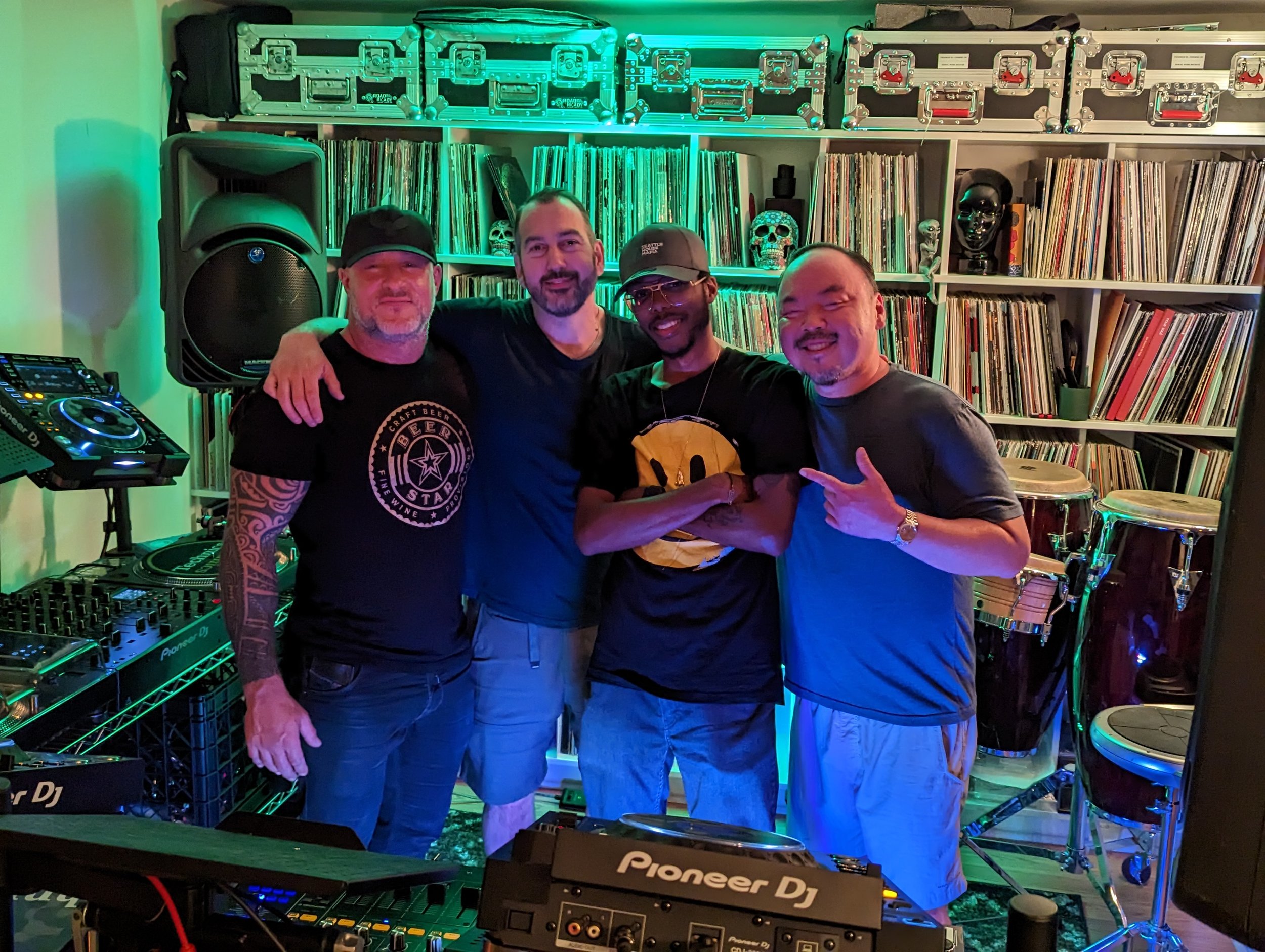 Phil Anthony, Paul Velocity, Tony H and Sang-Do posing together in a home studio with Pioneer DJ equipment, vinyl record collection, and green ambient lighting.