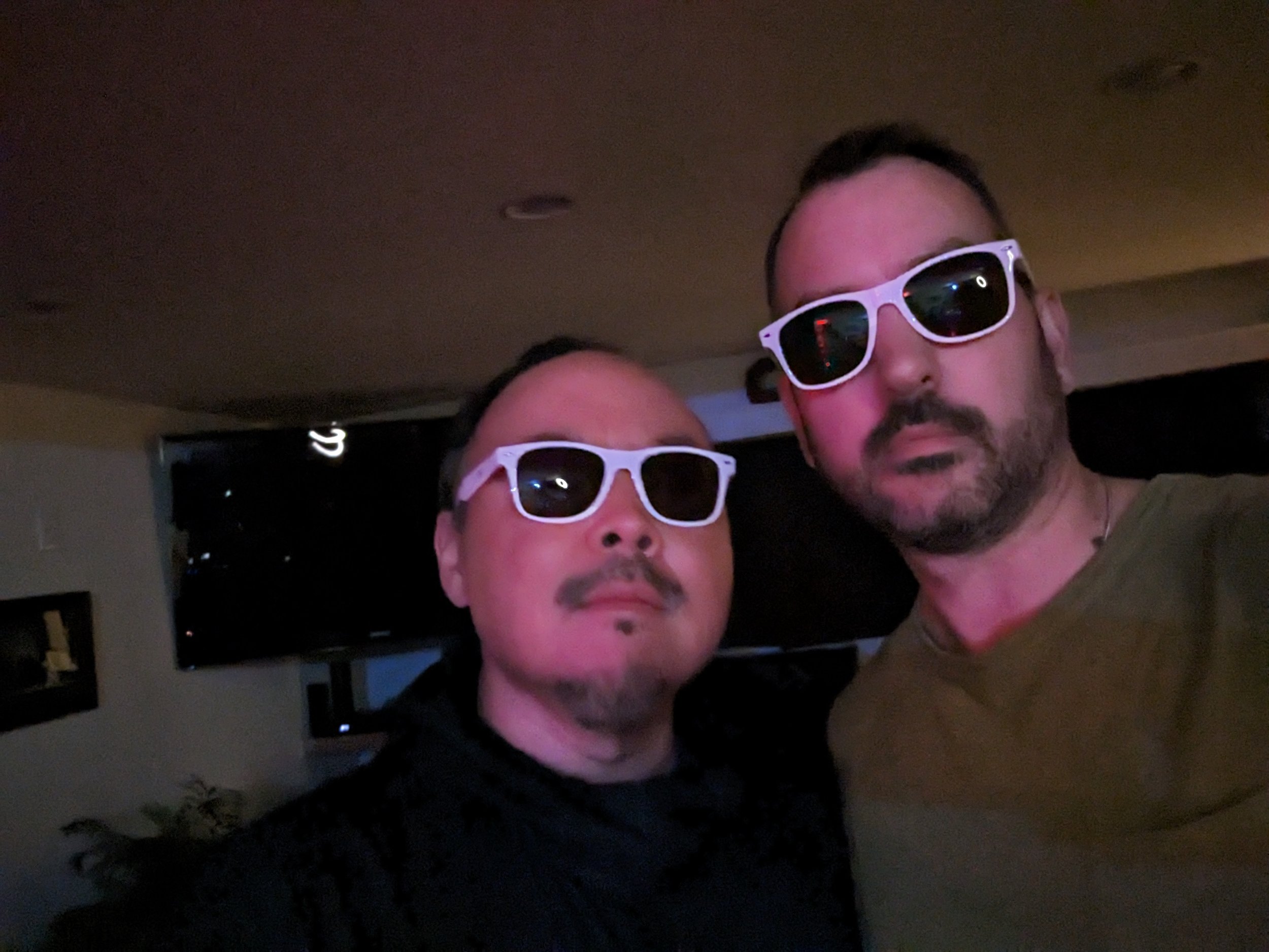 Sang-Do and Paul Velocity taking a selfie together wearing matching white sunglasses in a dimly lit room.