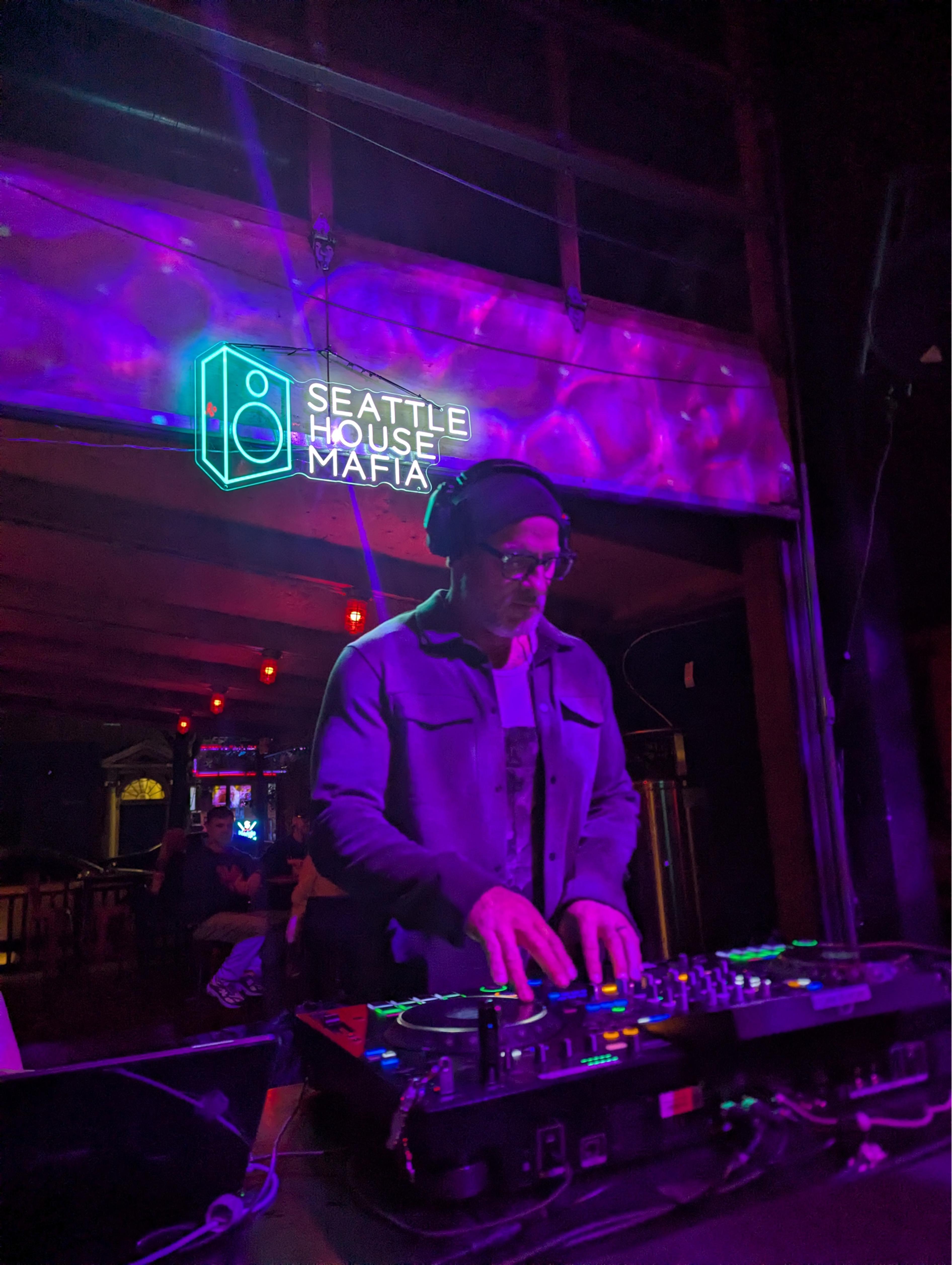 DJ mixing beneath the Seattle House Mafia neon sign under purple stage lighting