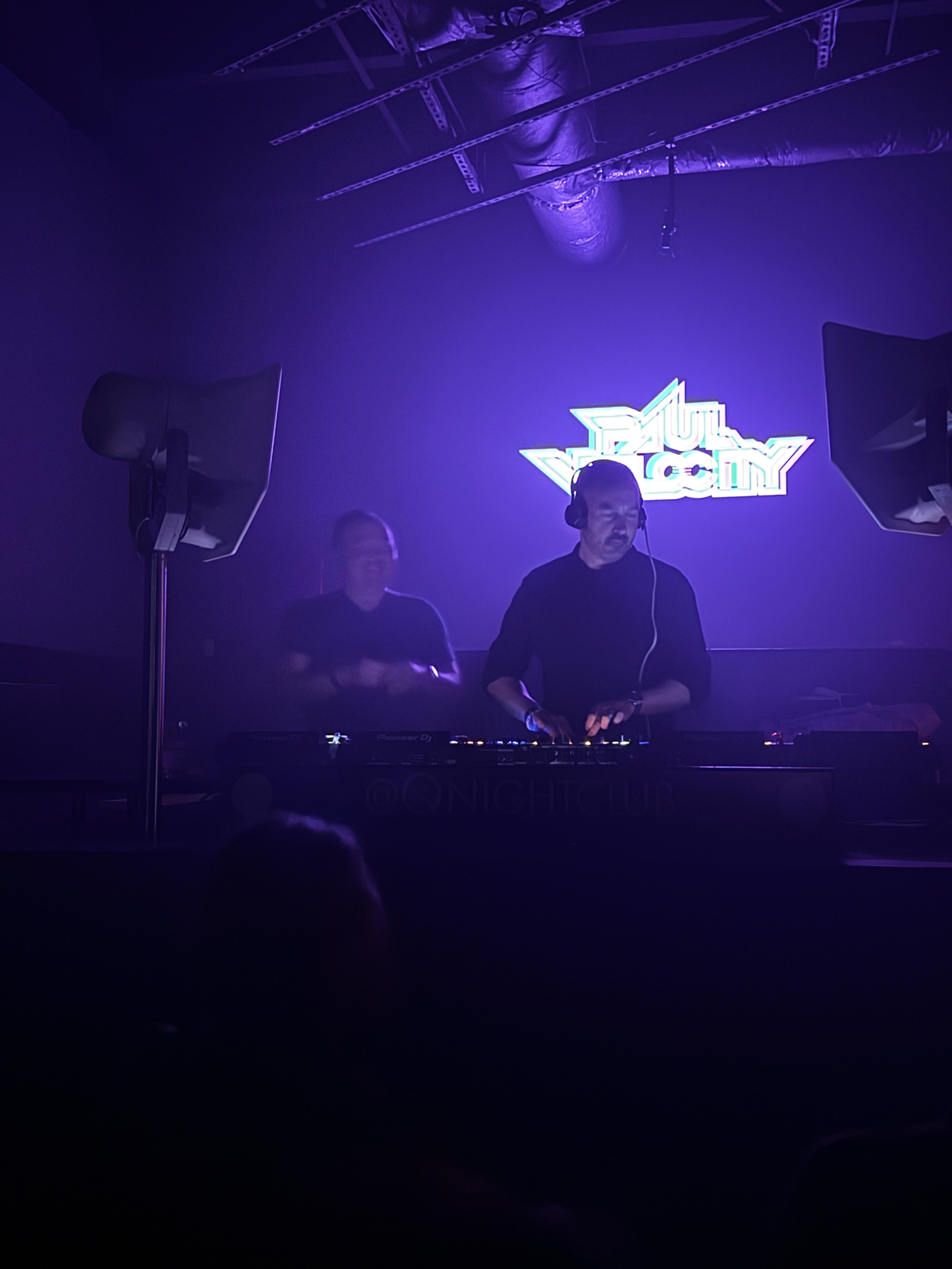 Two DJs behind the decks beneath the Paul Velocity neon sign at Q Nightclub