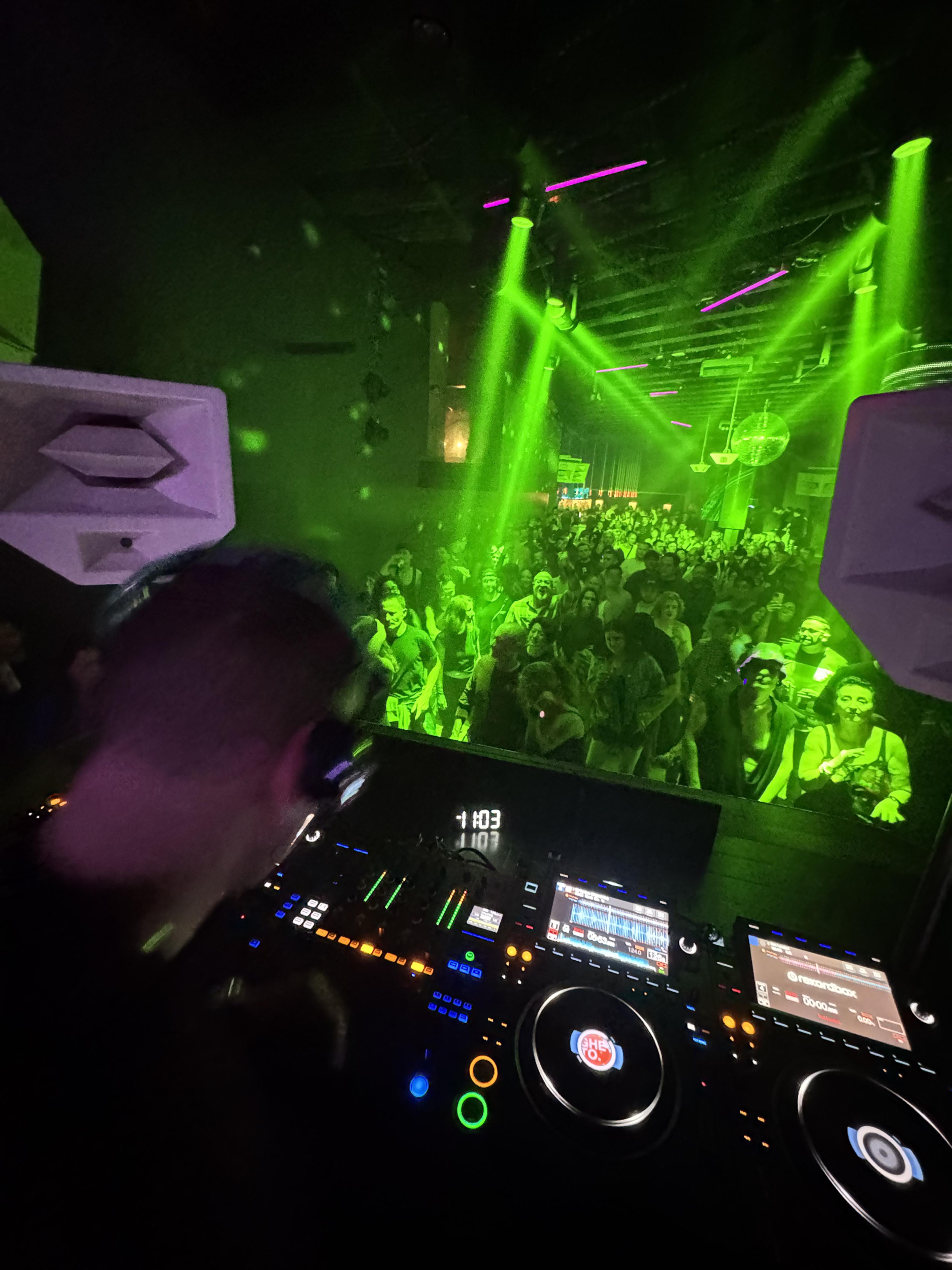 Green laser beams sweeping over a crowded dance floor, seen from behind the CDJs