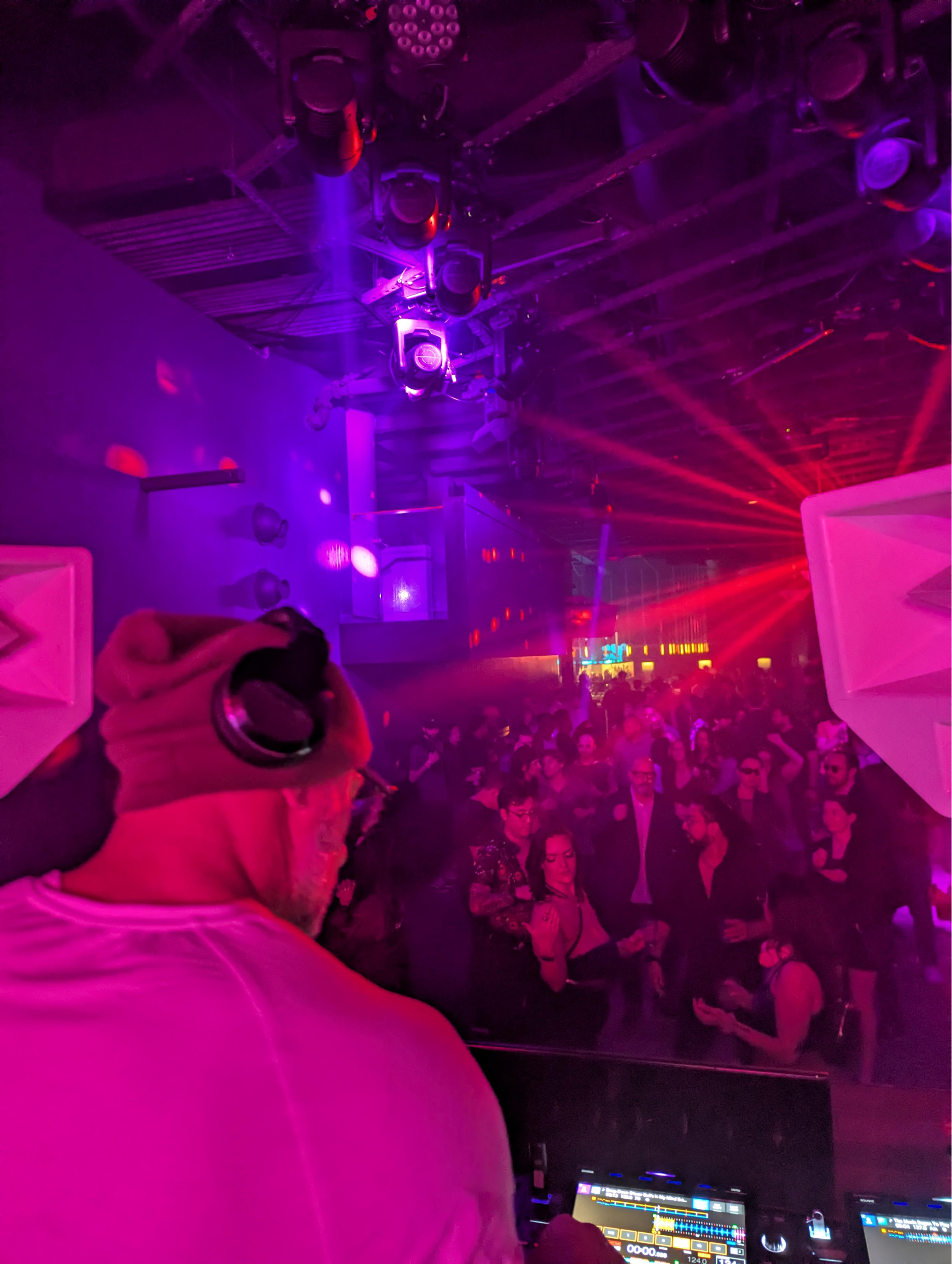 View from behind the DJ booth across a packed dance floor bathed in red and magenta light