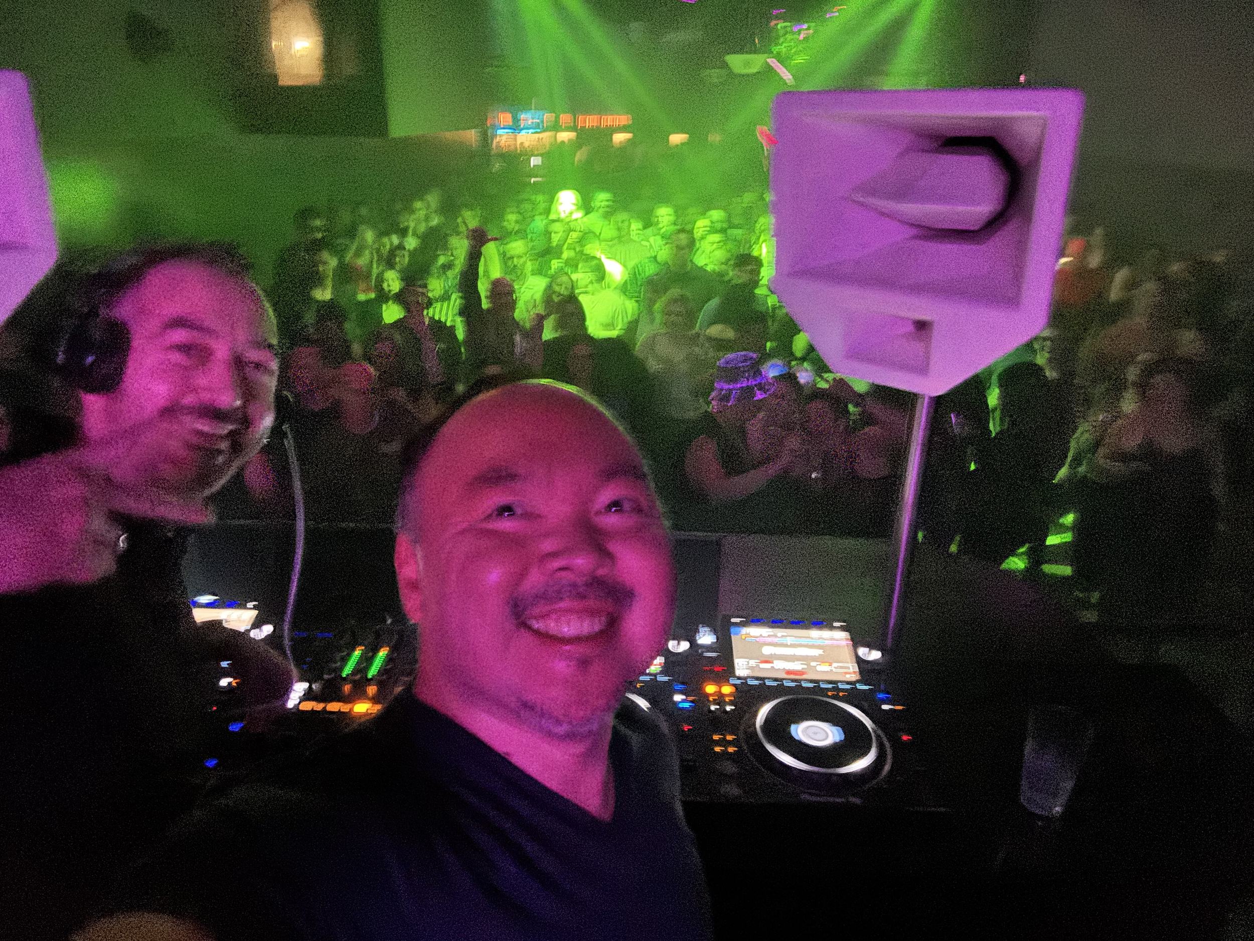 Two Seattle House Mafia DJs in the booth, one waving to a green-lit crowd behind them